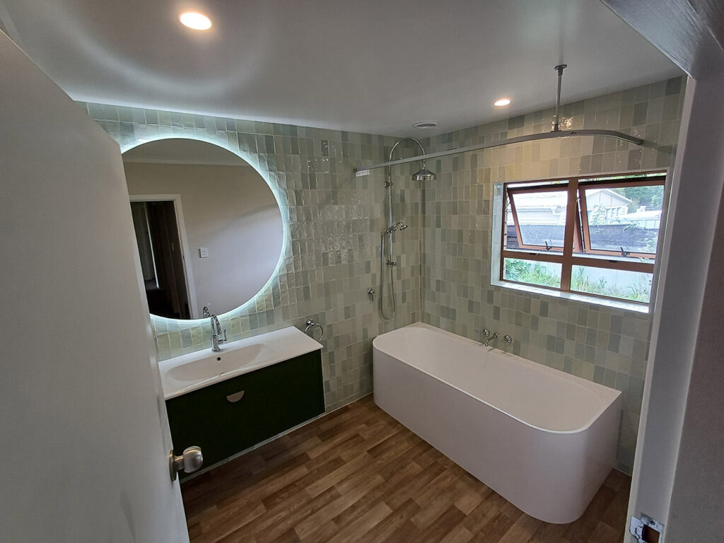 Wide view of a green tiled bathroom with freestanding bath and LED mirror